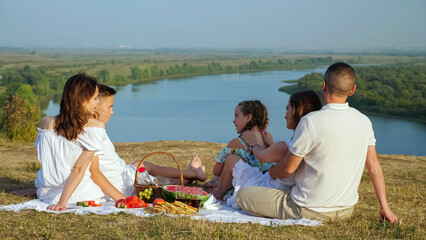 Large family with children of different ages enjoys picturesque river view from green hilly riverbank at festive picnic on sunny summer day backside view