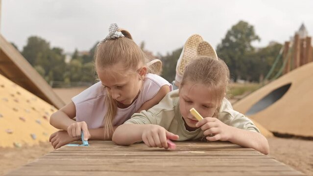 Slowmo Of Two Pretty 10 Year Old Caucasian Girls Drawing With Chalk While Lying On Wooden Bench At Playground On Summer Day