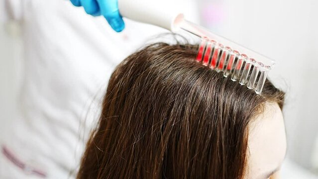 Professional beautician doing darsonval hair treatment procedure for young woman in beauty salon