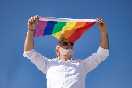 Mature, gray-haired gay man with beard, sunglasses and white shirt waving a gay pride flag in the wind under a blue sky. Concept mature gay man, sugar daddy, lgbt, lgbtiq+, pride, teddy bear.