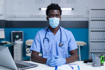 Hospital medical staff sitting at table in clinic cabinet while wearing surgical gloves and virus protection mask. Healthcare nurse sitting at office desk while wearing facemask.