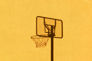 Der Schatten eines Basketballkorbes zeichent sich durch das Sonnenlicht auf einer gelben Hauswand ab