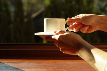 Female hands hold cup of hot drink near windowsill