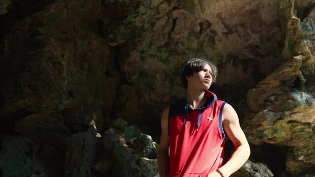 Young Asian man solo travel in rock mountain cave on tropical island beach lagoon in summer sunny day. Happy guy relax and enjoy outdoor activity lifestyle on summer holiday vacation trip in Thailand