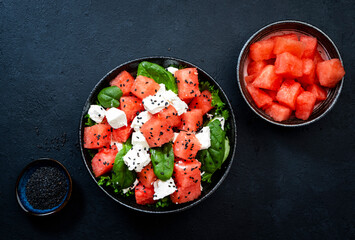 Summer Watermelon juicy salad bowl with feta cheese, spinach and black sesame seed, dark table background, top view, negative space