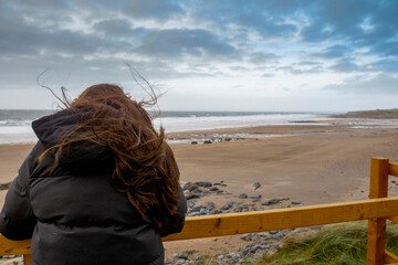 Teenager standing by wooden barrier and looking at beautiful Fanore beach. Ireland. Blue cloudy sky and Atlantic ocean. Travel and tourism concept.
