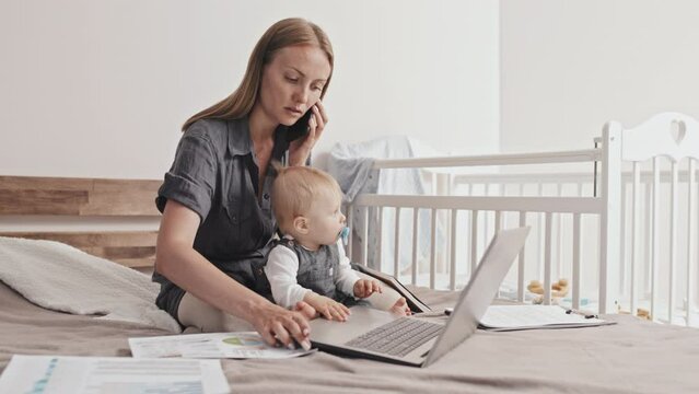 Full shot of young Caucasian woman sitting on bed with her toddler son, working with documents and portable computer, talking on mobile phone