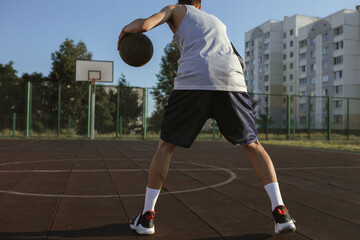 Back view of sportsman playing basketball alone on sports ground in summer. Man playing street ball on sunny day on basketball court 