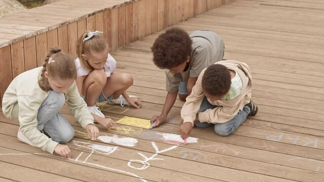 Slowmo of four multiethnic children talking while drawing with chalk on wooden planks at playground in summer