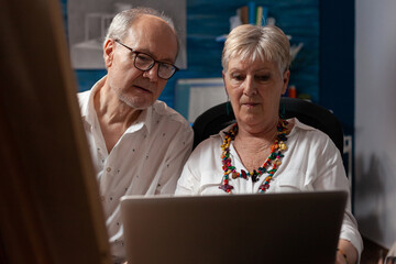 Fototapeta premium Elderly artist couple holding laptop looking at screen trying to sell artwork in online auction sitting in home studio. Senior man and woman art creators browsing social media for inspiration.