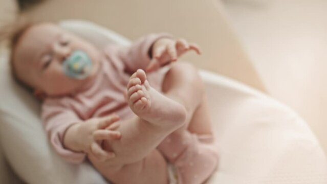 Selective Focus Of Feet Up Of Cute Blue-eyed Biracial Infant In Pink Onesie With Pacifier Lying In Baby Nest On Couch At Home