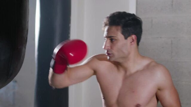 Strong male boxer hitting punching bag with force while working out in boxing gym alone. Attractive young man training indoors. Handheld shot. Slow motion. Sport, boxing concept