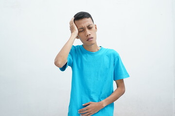 Portrait of young man wearing blue t-shirt isolated on white background suffering from severe headache, closing eyes to relieve pain with helpless facial expression