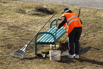 European janitor worker woman cleans the grass with a broom near a bench at spring day