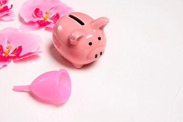 Menstrual cup with piggy bank and orchids on a white background. Save money with reusable feminine hygiene products.