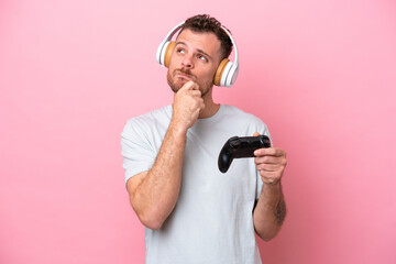 Young Brazilian man playing with video game controller isolated on pink background having doubts and thinking