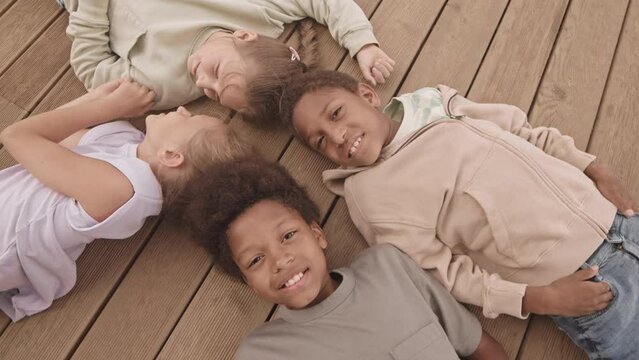 From above slowmo of four cheerful diverse kids smiling and waving at camera while lying on back together on wooden floor outdoors in summer