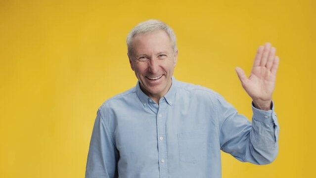 Hello. Happy excited sociable senior man waving hand to camera and smiling, greeting cheerfully, yellow background