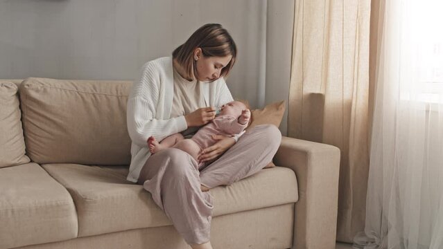Young short-haired Biracial woman sitting on couch in living room at daytime, holding her crying baby daughter, giving her pacifier