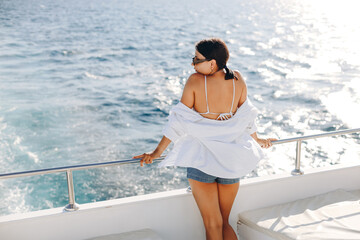 Stylish graceful woman with sun glasses looking at seasides resting on luxury yacht in summer day....
