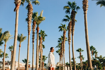 A beautiful young happy girl enjoys a sunny day against the background of palm trees and blue sky. Horizontal photo