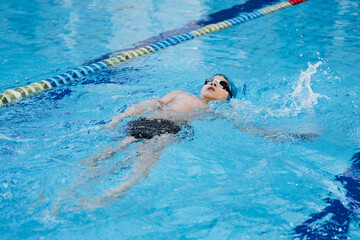 little caucasian boy wearing goggles swimming the backstroke in a pool