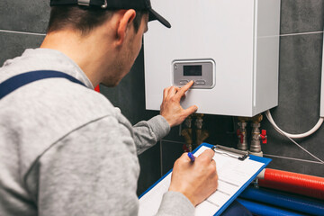 Technician servicing holding clipboard and inspecting heating system in boiler room - obrazy, fototapety, plakaty