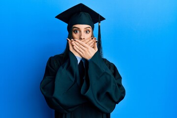 Beautiful brunette young woman wearing graduation cap and ceremony robe shocked covering mouth with...