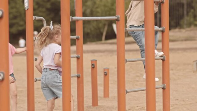 Slowmo of pretty Caucasian girl climbing up and tumbling on playground facility with her multiethnic schoolfriends, spending summertime together outdoors