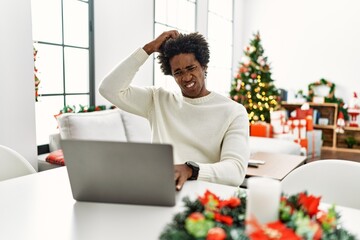 Young african american man using laptop sitting on the table by christmas tree confuse and wonder about question. uncertain with doubt, thinking with hand on head. pensive concept.