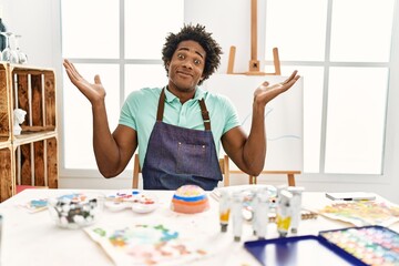 Young african american man sitting on the table at art studio clueless and confused expression with arms and hands raised. doubt concept.