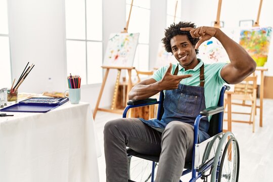 Young african american artist man sitting on wheelchair at art studio smiling making frame with hands and fingers with happy face. creativity and photography concept.