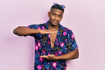 Young black man wearing hawaiian shirt and sunglasses gesturing with hands showing big and large size sign, measure symbol. smiling looking at the camera. measuring concept.