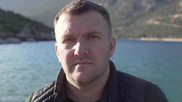 Close-up, a serious man stands by the sea and looks at the camera.