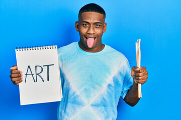 Young black man holding art notebook and painter brushes sticking tongue out happy with funny expression.