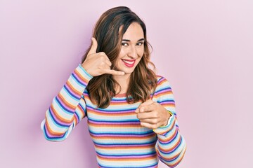 Young hispanic girl wearing casual clothes smiling doing talking on the telephone gesture and pointing to you. call me.