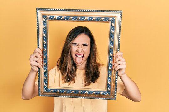 Young hispanic girl holding empty frame sticking tongue out happy with funny expression.