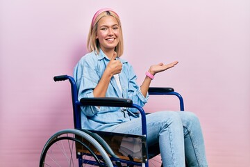 Beautiful blonde woman sitting on wheelchair showing palm hand and doing ok gesture with thumbs up,...