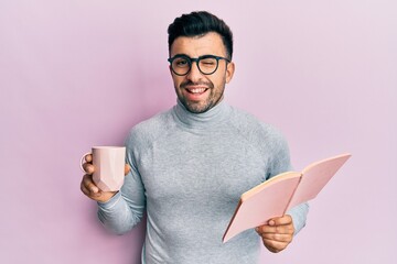 Young hispanic man reading a book and drinking a cup of coffee winking looking at the camera with...