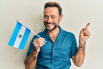 Middle age man holding argentina flag smiling happy pointing with hand and finger to the side - obrazy, fototapety, plakaty
