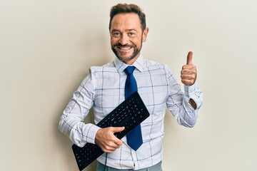 Middle age businessman holding keyboard smiling happy and positive, thumb up doing excellent and approval sign