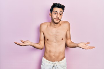 Fototapeta premium Young hispanic man wearing swimwear shirtless clueless and confused expression with arms and hands raised. doubt concept.