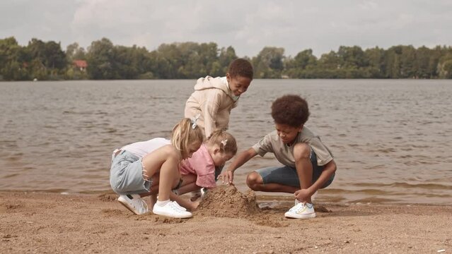 Slowmo of four cheerful multiethnic kids building sandcastle on beach by lake in summertime