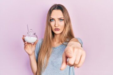 Young blonde girl holding bowl with sugar pointing with finger to the camera and to you, confident gesture looking serious