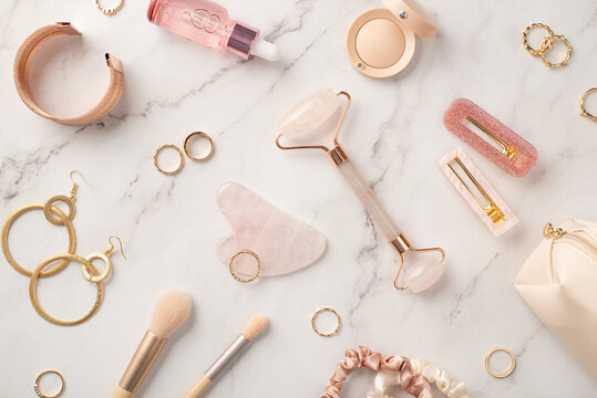 Top view photo of pink barrettes rose quartz roller gua sha dropper bottle cosmetics eyeshadow scrunchies makeup brushes golden earrings rings wristlet and cosmetic bag on white marble background