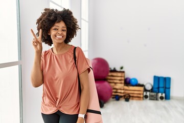 Obraz na płótnie Canvas African american woman with afro hair holding yoga mat at pilates room smiling looking to the camera showing fingers doing victory sign. number two.