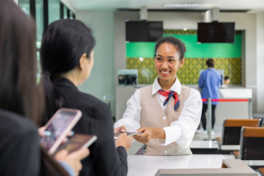 Smiling multiracial woman in check-in counter taking passport for verification from passenger. People waiting in queue at airplane counter at airport..
