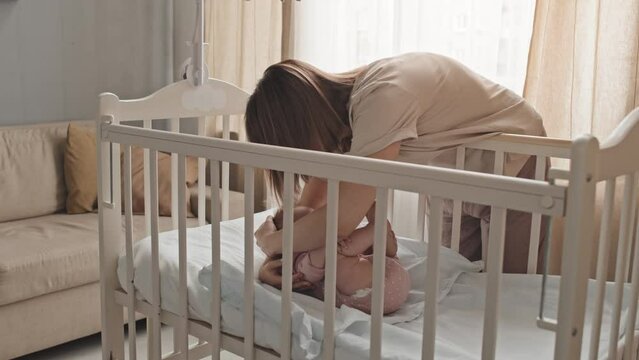 Medium long of young brown-haired woman putting sleeping baby girl with pacifier into crib at home in afternoon