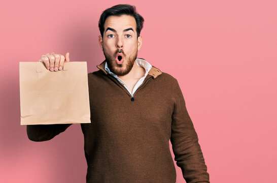 Young hispanic man holding take away paper bag scared and amazed with open mouth for surprise, disbelief face