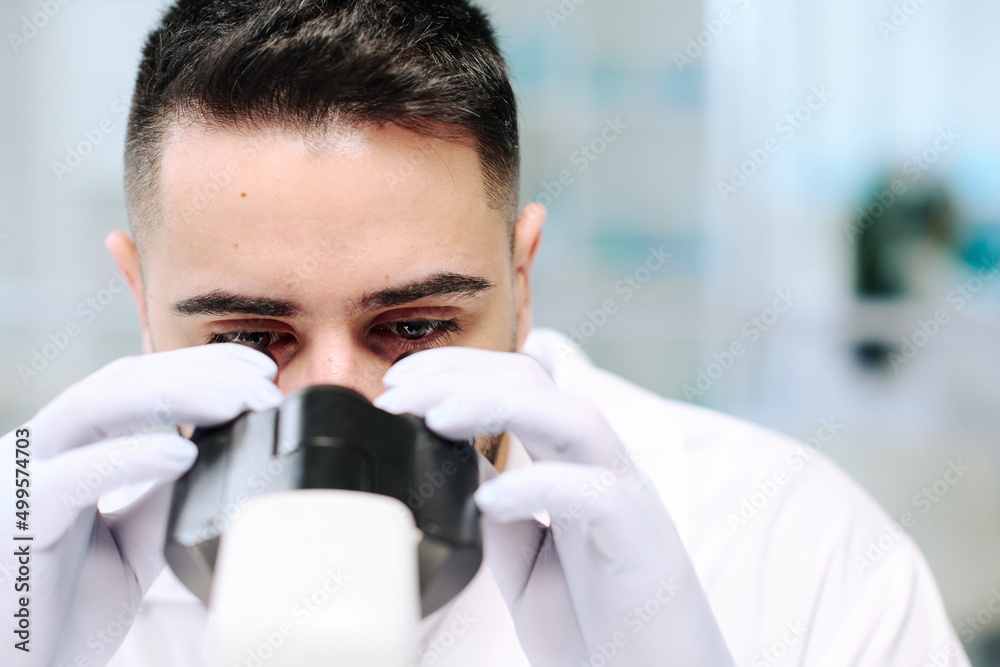 Poster close-up of face of young serious male scientist in gloves looking in microscope during scientific e